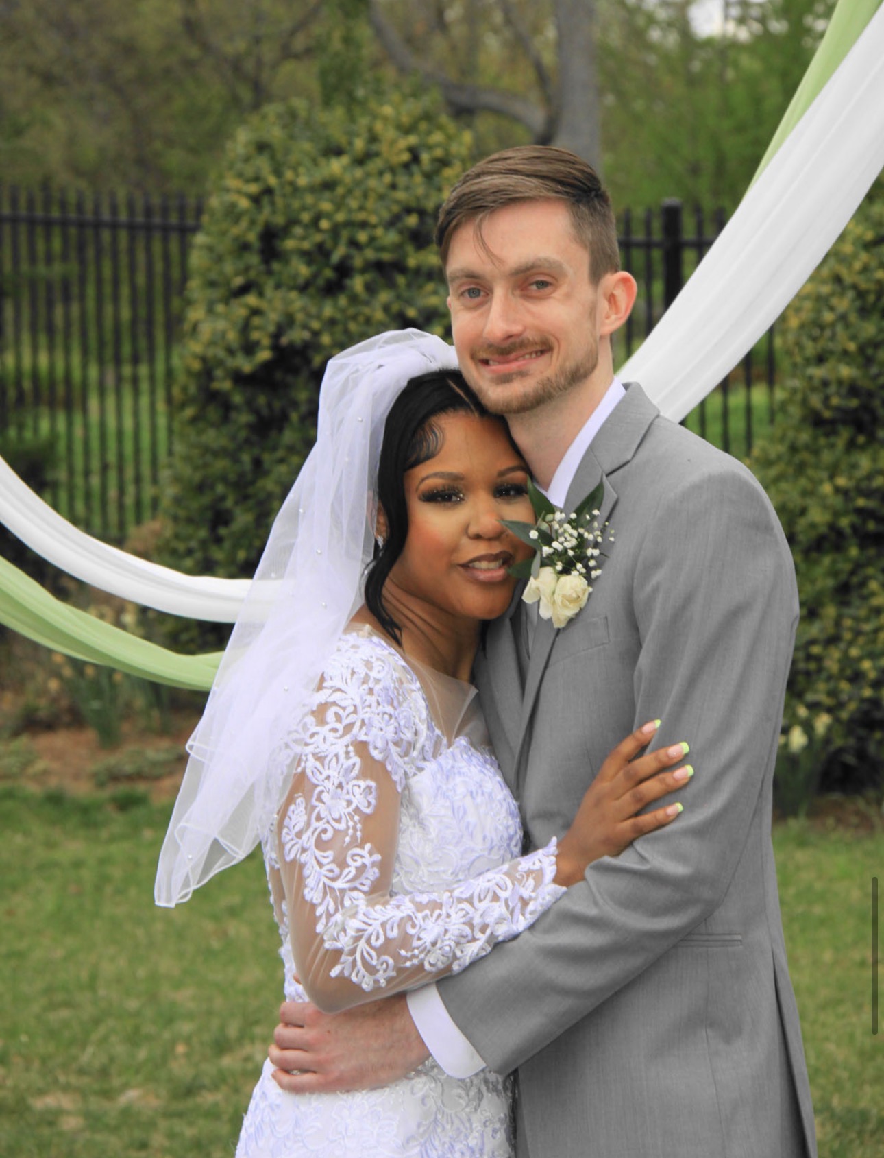 Bride in lace sleeves and groom in a grey suit embracing beneath a draped garden arch