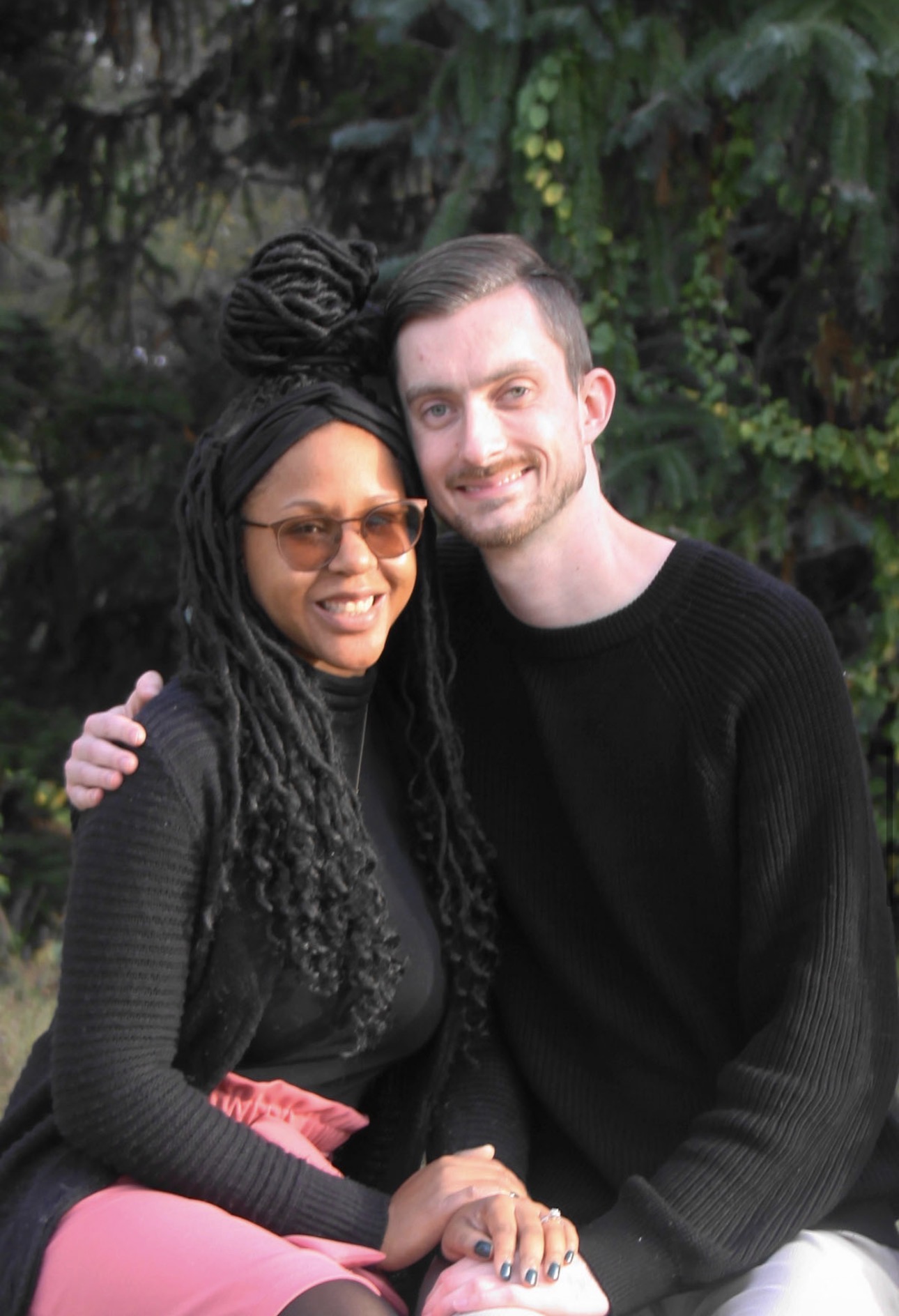 Engaged couple seated close together in front of evergreen branches, showing the ring